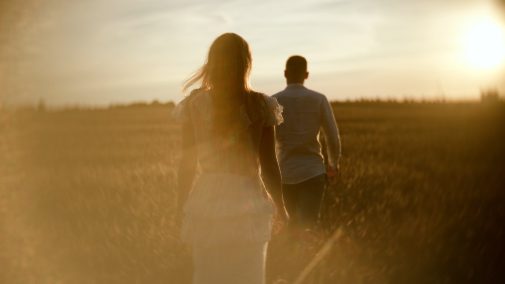 Klatka z filmu plenerowego "Your Story", para idzie przez pole w stronę zachodzącego słońca.